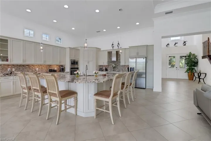 Kitchen featuring wall chimney range hood, appliances with stainless steel finishes, a kitchen island with sink, light stone countertops, and pendant lighting