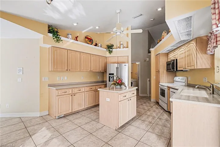 Kitchen with light brown cabinetry, high vaulted ceiling, 2 appliances with stainless steel finishes, , 2 appliancies with white finishes, light solid surface countertops, and light tile patterned flooring