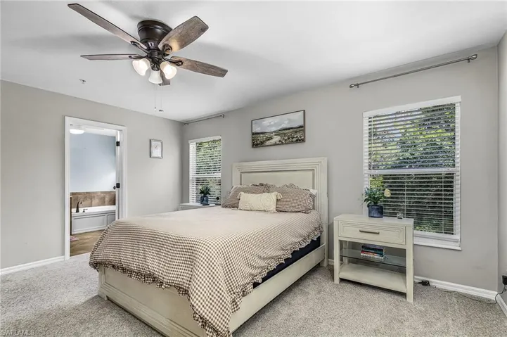 Carpeted bedroom featuring a dark bronze ceiling fan with light kit, two windows with blinds, and an open doorway revealing a bathroom with a built-in tub
