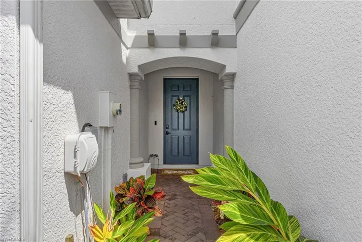 Arched portico entrance featuring a paneled door, decorative columns, textured exterior finish, and a brick paver walkway bordered by lush landscaping