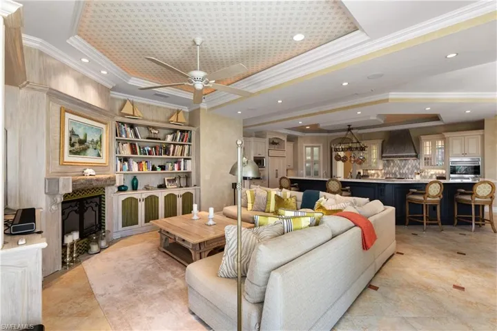 Tiled living room with a tray ceiling, a premium fireplace, crown molding, built in shelves, and ceiling fan with notable chandelier