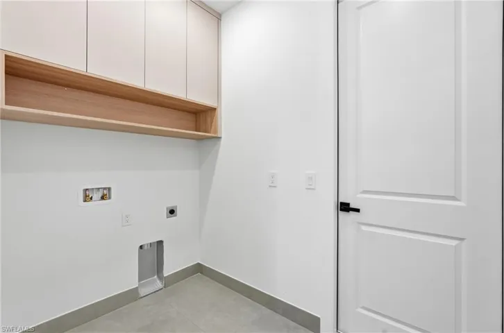 Dedicated laundry space featuring upper cabinetry, an open wood-finish shelf, and contemporary gray floor tiles