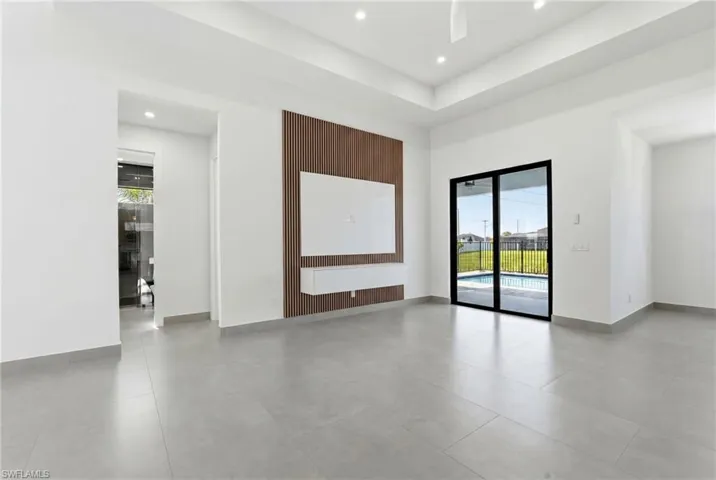Spacious interior featuring light gray tile flooring, a contemporary wood-slat accent wall with built-in media console, a tray ceiling with recessed lighting, and expansive glass sliding doors providing direct access to the backyard and pool