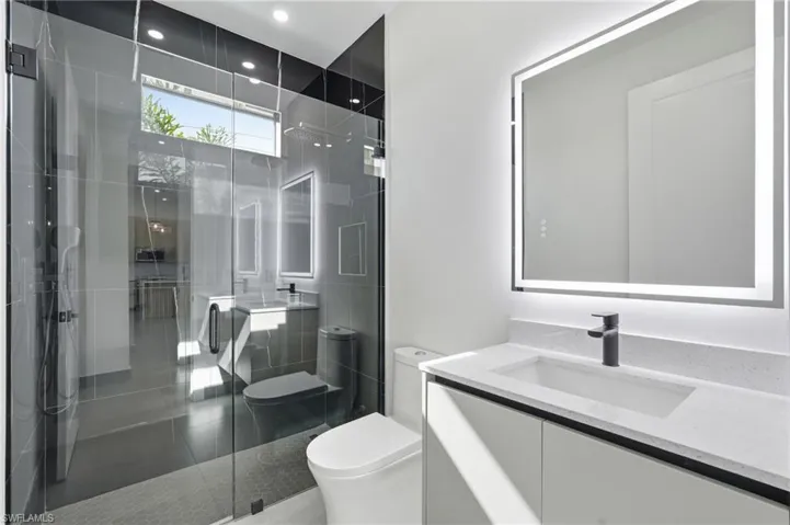 Contemporary bathroom featuring a frameless glass shower enclosure, a vanity with a white countertop and integrated sink, a backlit mirror, and dark-finish plumbing fixtures