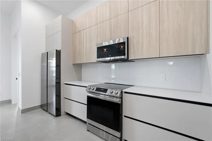 Contemporary kitchen featuring wood-finish upper cabinetry, white lower cabinetry with black trim, stainless steel appliances, a white speckled backsplash, and white countertops