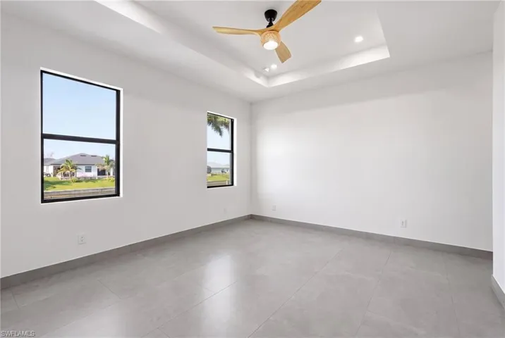 Spacious room featuring light gray tile flooring, crisp white walls, two large black-framed windows, a decorative tray ceiling with recessed lighting, and a modern ceiling fan