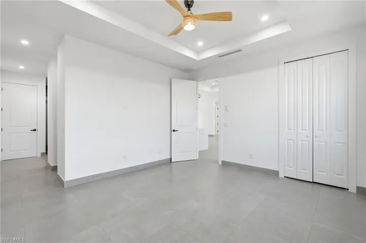 Spacious room featuring light gray tile flooring, crisp white walls, a tray ceiling with recessed lighting, a ceiling fan with wood-tone blades, and double bifold closet doors