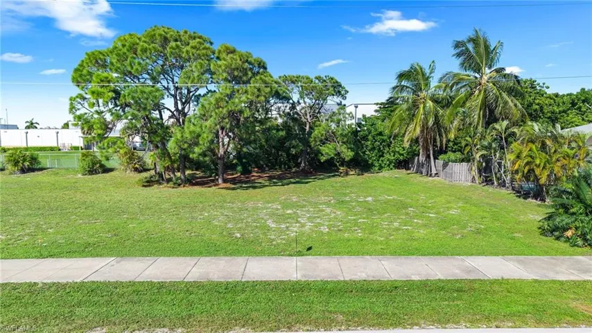 Vacant parcel featuring expansive grass lawn, mature pine trees, palm trees, and concrete sidewalk frontage