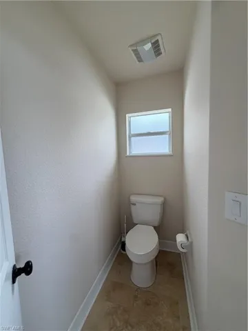 Compact powder room featuring a window, ceiling-mounted exhaust fan, tiled flooring, travertine tiles, white baseboards, and a white toilet