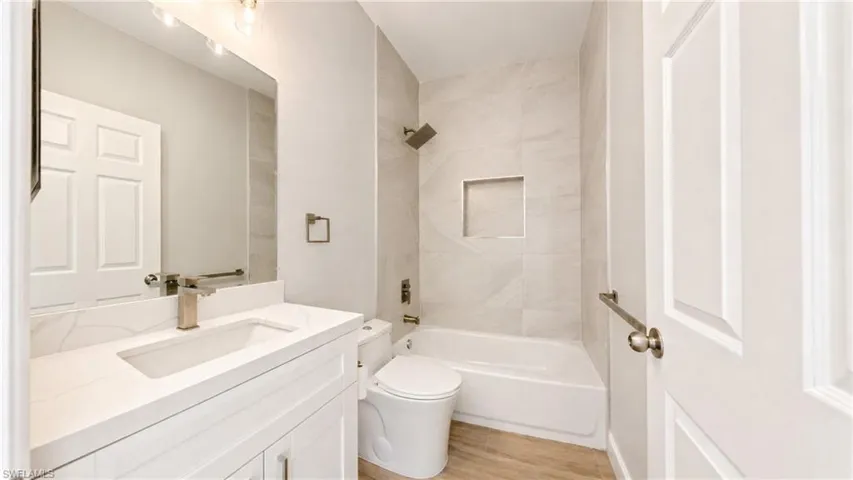 Bathroom featuring a white vanity with an integrated rectangular sink and gold-tone faucet, a white bathtub, a tiled shower surround with a recessed niche, and wood-finish flooring