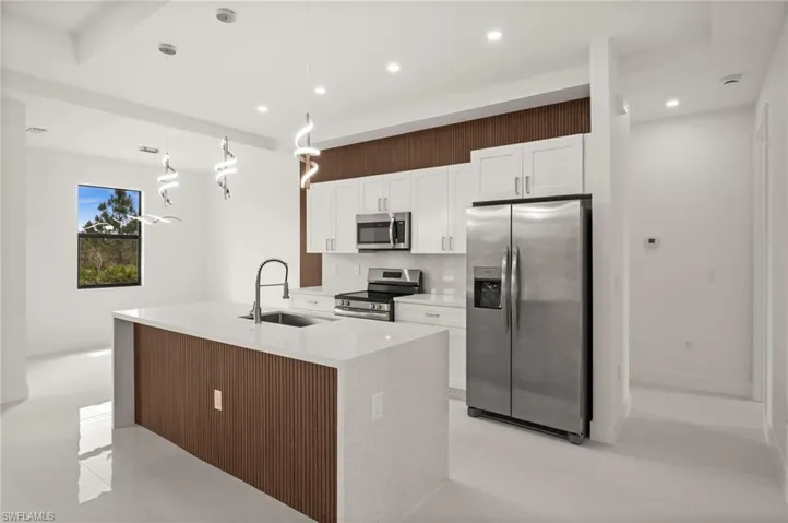 Kitchen with stainless steel appliances, light stone counters, a center island with sink, white cabinetry, and decorative light fixtures