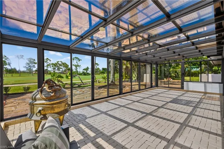 Spacious sunroom featuring a clear glass and metal frame ceiling, floor-to-ceiling glass panel walls, light-toned paver flooring, and white built-in cabinetry