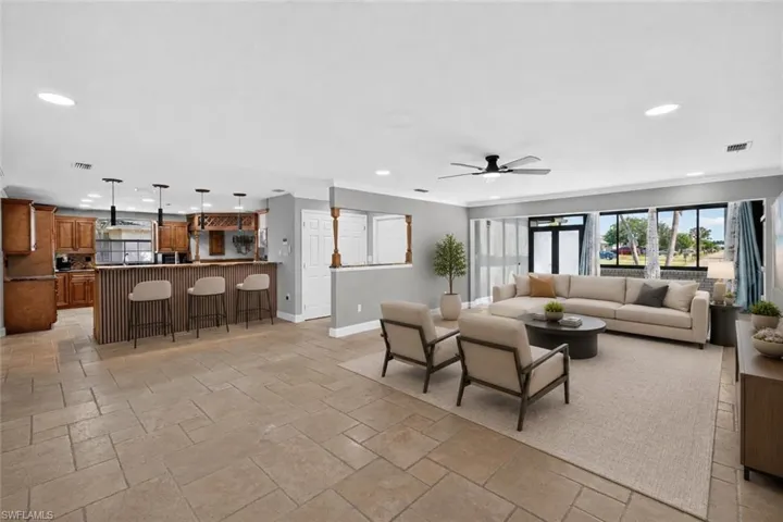 Open concept living area featuring light-toned tile flooring throughout, a kitchen with wood cabinetry, and a living room with multiple large windows, Virtually Staged