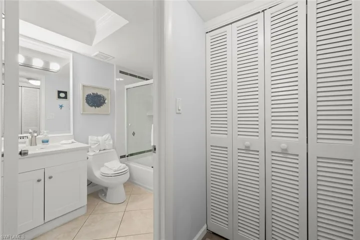 Bathroom featuring a white vanity cabinet with a rectangular undermount sink, tiled flooring, built-in louvered-door cabinetry, and a shower-tub combination with glass enclosure