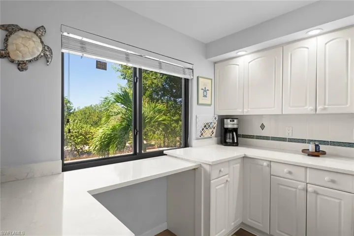 Kitchen featuring white cabinetry, light-toned stone-finish countertops, tile backsplash with decorative accent inlay, and a large window with exterior tropical greenery