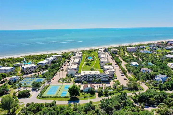 Aerial perspective of coastal residential property featuring multiple swimming pools, tennis courts, paved walkways, and direct proximity to the shoreline