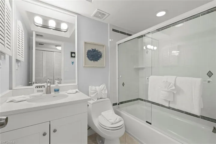 White vanity cabinet with an integrated sink and quartz countertop, complemented by a large-format mirror, tri-light fixture, and a tub-shower combination featuring a sliding glass enclosure with tile surround