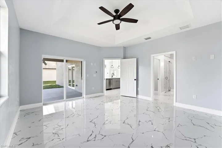 Unfurnished bedroom with ensuite bath, a tray ceiling, access to exterior, ceiling fan, and light marble finish flooring