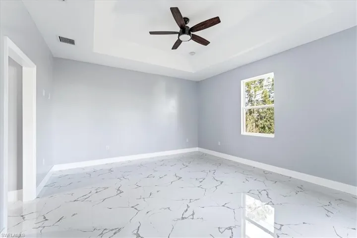 Empty room with a raised ceiling, light marble finish floors, and a ceiling fan