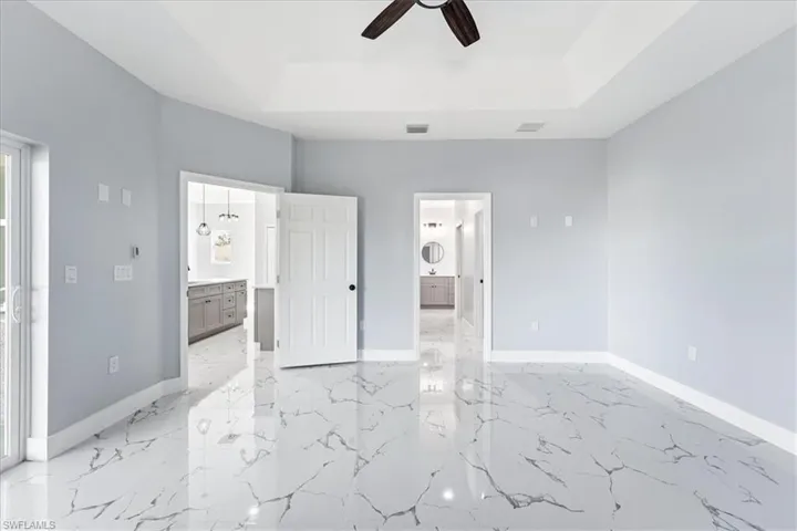 Unfurnished bedroom featuring ensuite bath, a tray ceiling, ceiling fan, and light marble finish floors