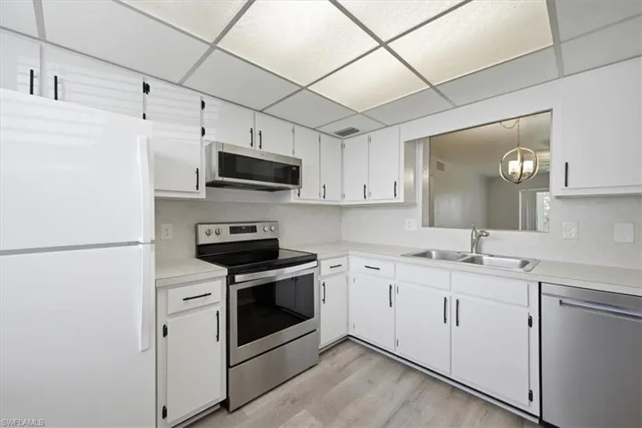 Kitchen featuring white cabinetry with black hardware, stainless steel appliances, light wood-finish flooring, a double basin sink, and a drop ceiling with integrated lighting