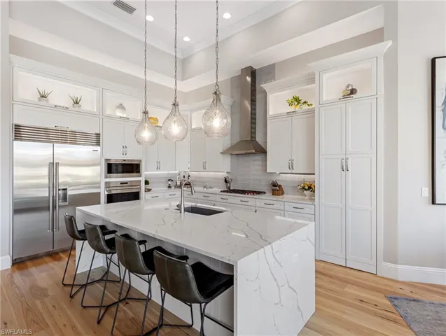 Kitchen featuring sink,  range hood, built in appliances, a kitchen island with sink, and white cabinets