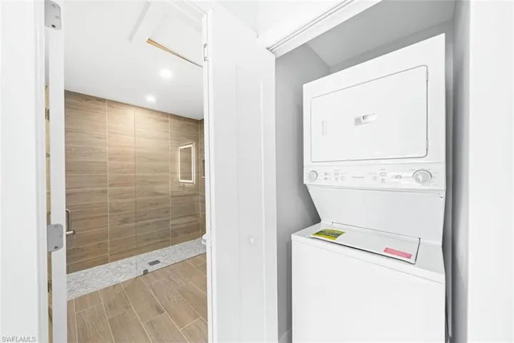 Stacked laundry unit housed in a dedicated closet, featuring walk-in shower with wood-finish tile wall surround, recessed lighting, and stone mosaic flooring