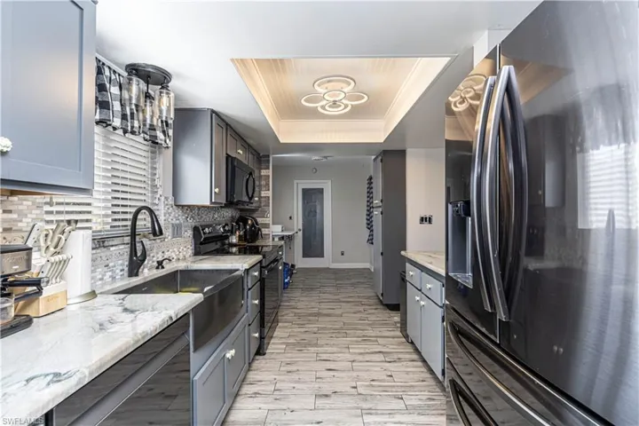Kitchen with backsplash, light stone countertops, light wood-type flooring, black appliances, and a raised ceiling
