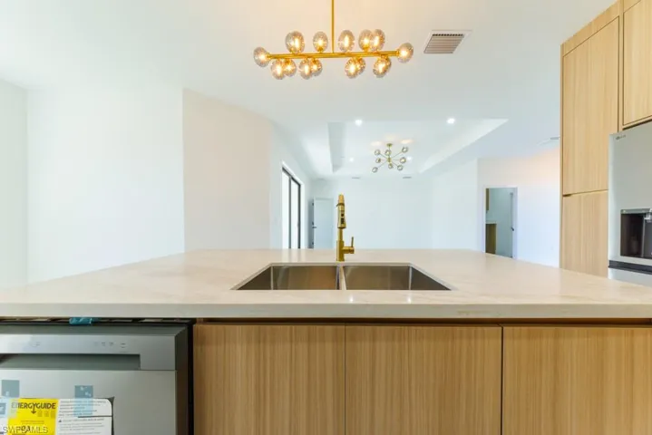 Kitchen featuring light brown cabinetry, dishwasher, a tray ceiling, light stone countertops, and a kitchen island with sink