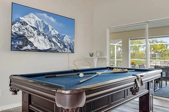 Open-concept room featuring a wall-mounted display, a dark wood-finish billiards table with blue felt, and large sliding glass doors leading to an outdoor area