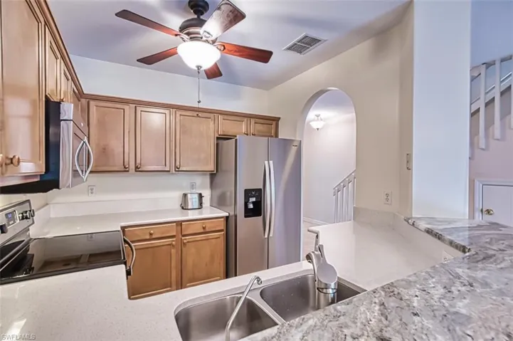 Kitchen with appliances with stainless steel finishes, ceiling fan, arched walkways, brown cabinets, and marble/quartz counters