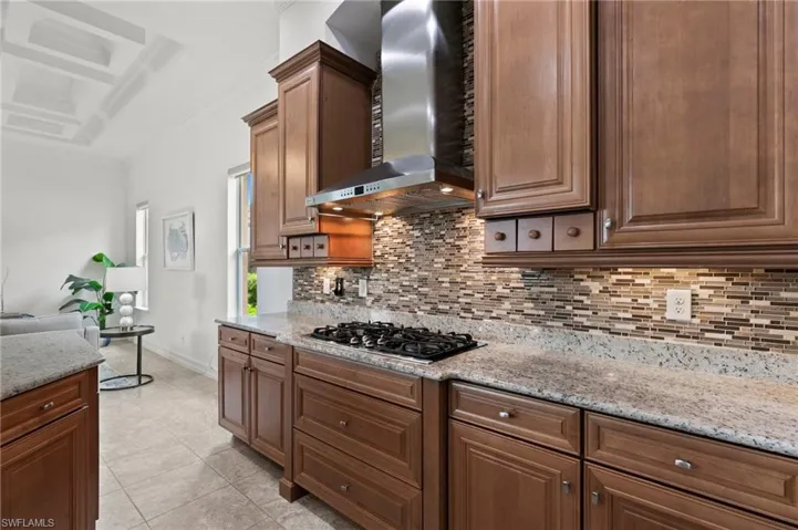 Kitchen with wall chimney range hood, gas stovetop, backsplash, light stone counters, and coffered ceiling