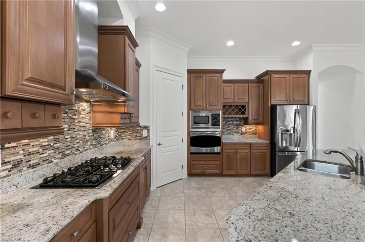 Kitchen featuring wall chimney exhaust hood, appliances with stainless steel finishes, tasteful backsplash, light stone counters, and crown molding