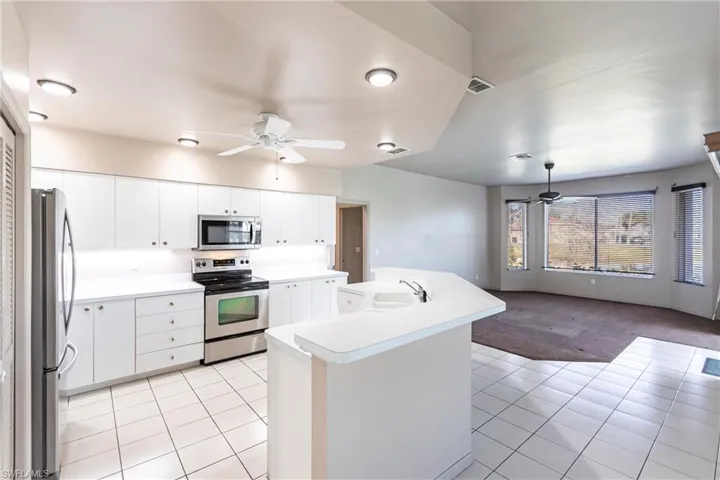 Kitchen with breakfast bar & dining area, white cabinetry, sink, light tile patterned floors, ceiling fan, and stainless steel appliances. View of carpeted entertainment area overlooking lake with bay window (manual shutters on all 3 windows), custom blinds, too!