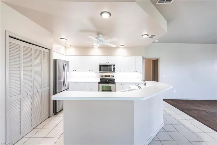 Kitchen with white cabinetry, spacious pantry, appliances with stainless steel finishes, a kitchen island with sink, and light tile patterned floors leading to carpeted entertainment area overlooking lake.