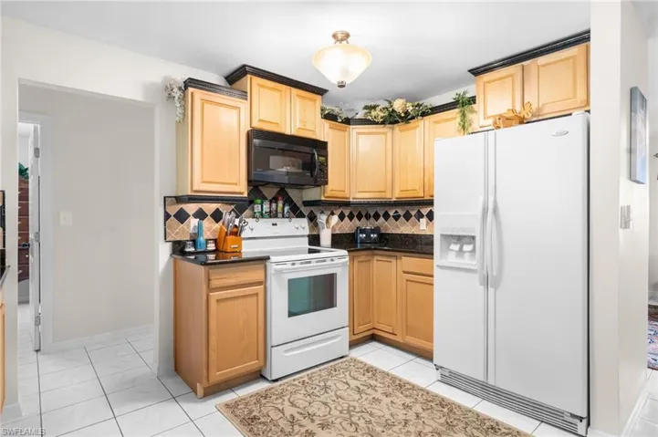 Kitchen featuring light wood cabinetry, dark countertops, a black over-the-range microwave, and white appliances