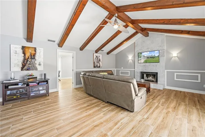 Vaulted ceiling with exposed wood beams, wood-finish flooring, accent fireplace with stone surround, wainscoting, and recessed lighting