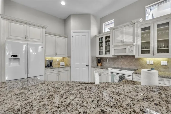Kitchen featuring white appliances, light stone countertops, glass insert cabinets, white cabinets, and tasteful backsplash