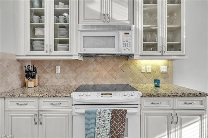 Kitchen with white appliances, glass insert cabinets, white cabinetry, light stone counters, and decorative backsplash