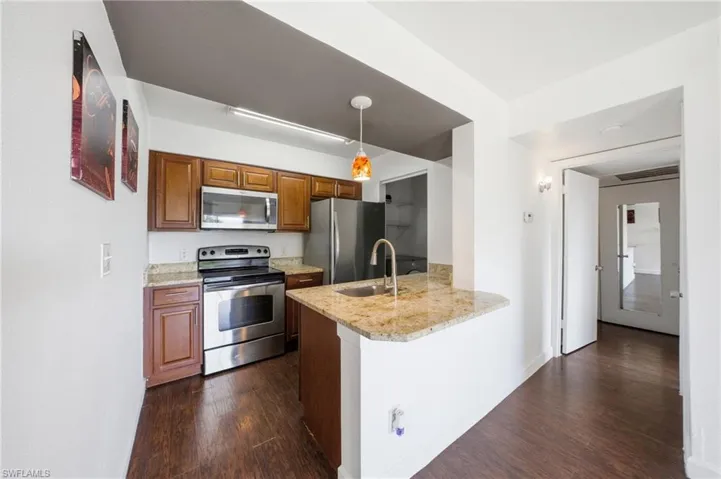 Kitchen with appliances with stainless steel finishes, light stone counters, pendant lighting, dark wood-style flooring, and brown cabinetry