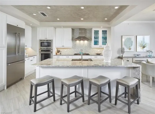 Kitchen featuring light stone countertops, built in appliances, decorative backsplash, white cabinetry, and a kitchen bar
