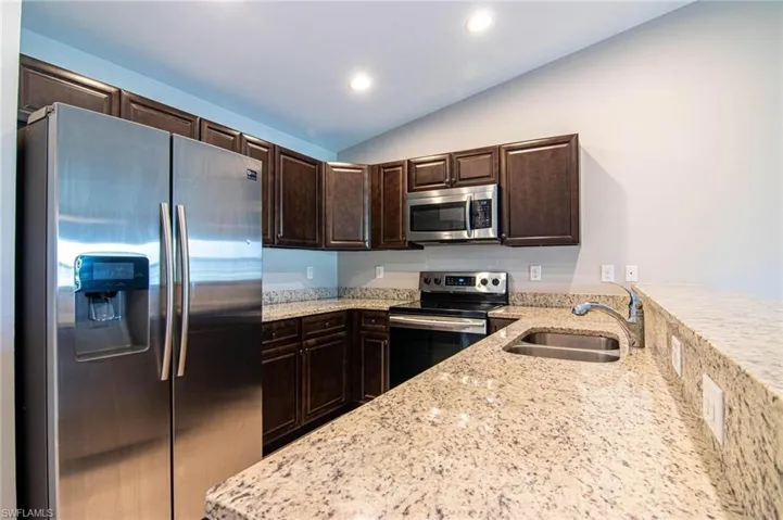Kitchen with stainless steel appliances, light stone counters, dark wood finish cabinets, vaulted ceiling, and a peninsula
