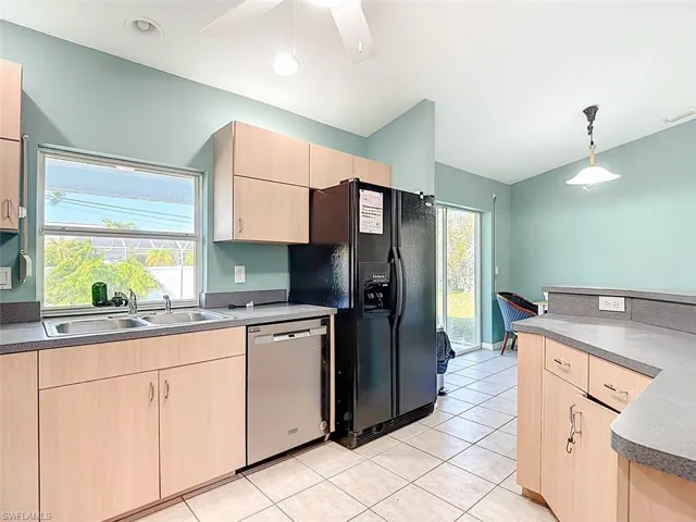 Kitchen with light brown cabinets, decorative light fixtures, stainless steel dishwasher, light tile patterned floors, and healthy amount of natural light
