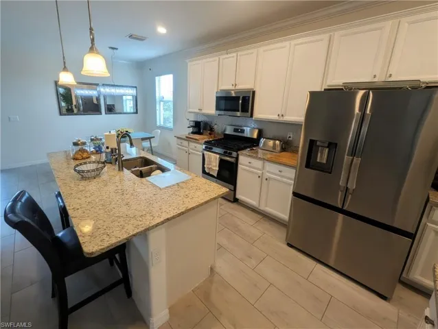 Modern kitchen featuring an island with a built-in sink, pendant lighting, stainless steel appliances, white cabinetry, and wood-finish tile flooring
