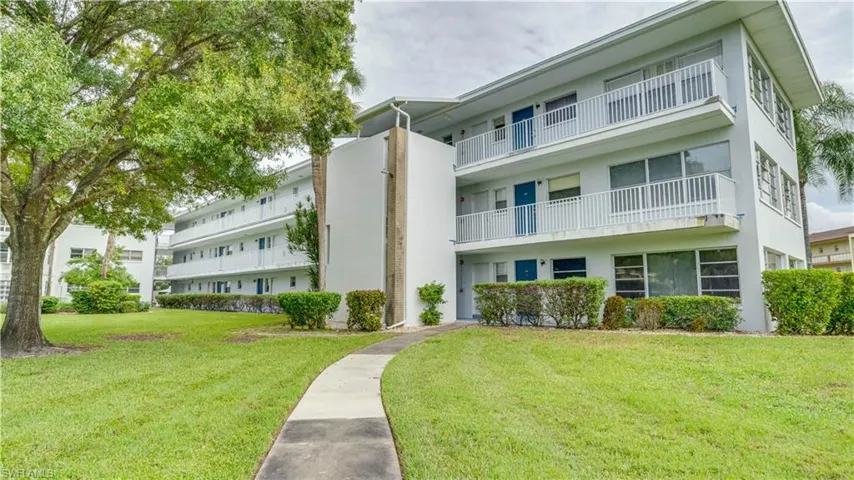 Three-story residential building with white exterior, multiple balconies with white railings, mature trees, and a curved sidewalk