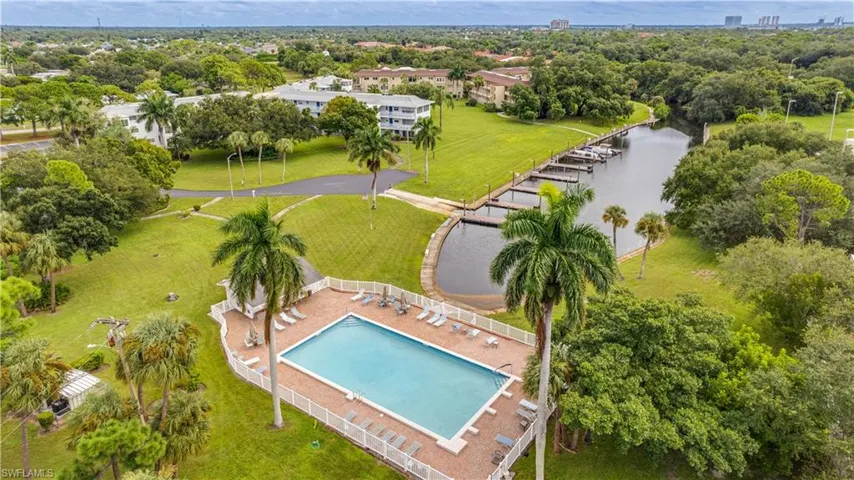 Community pool with a white fence enclosure, surrounded by expansive green spaces, mature palm trees, and a canal with boat docks