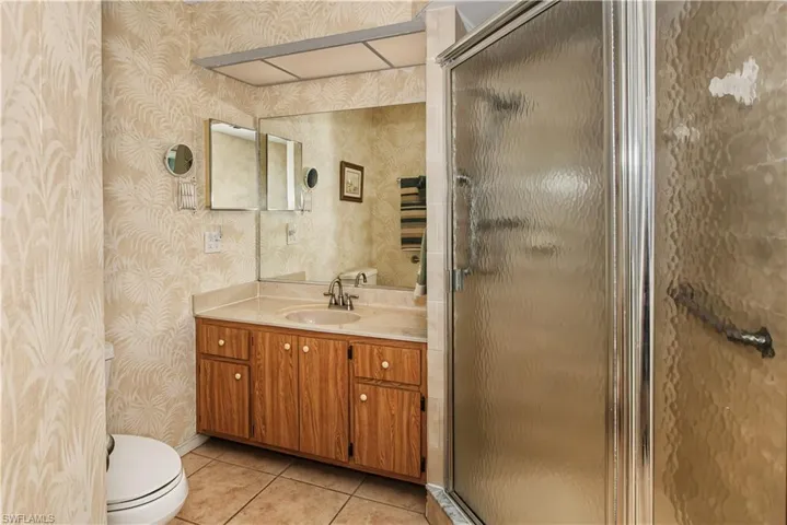 Spa-inspired primary bathroom featuring warm travertine tile walls, wood-finish vanity cabinetry with dual sink configuration, framed mirror with integrated lighting, and a frameless glass-enclosed shower enclosure with rainfall showerhead and built-in ceiling exhaust ventilation