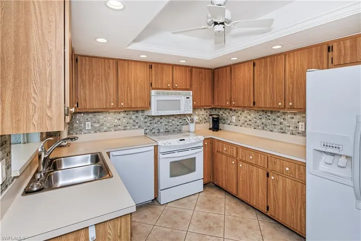 Warm wood cabinetry with stainless steel hardware frames this kitchen, featuring white appliances, ceramic tile flooring, and a mosaic tile backsplash