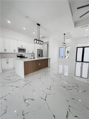 Open-concept interior featuring large format marble-look tile flooring, a kitchen island with a slatted wood-finish base, white shaker cabinetry, stainless steel appliances, and contemporary pendant lighting