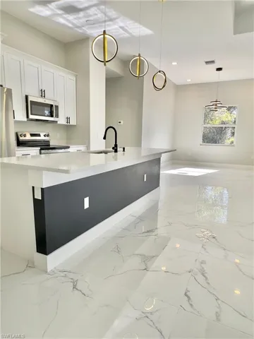 Kitchen featuring hanging light fixtures, appliances with stainless steel finishes, white cabinetry, light marble finish flooring, and a kitchen island with sink